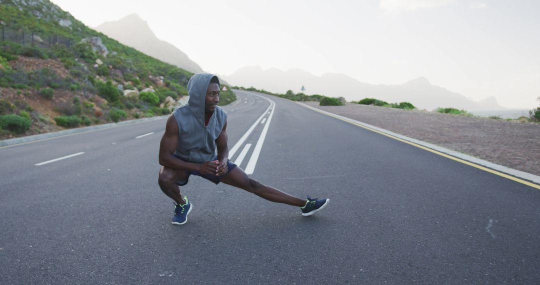 African american man wearing a hoodie performing stretching exercise on the road - Free Images, Stock Photos and Pictures on Pikwizard.com