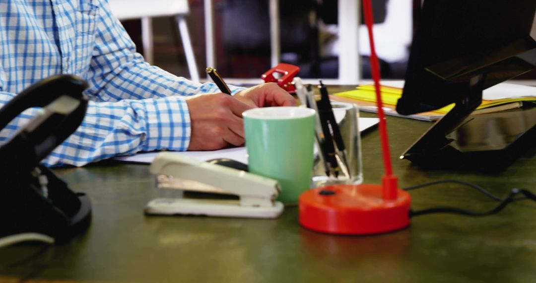 Man writing at office desk with stapler coffee mug and red lamp, workspace productivity