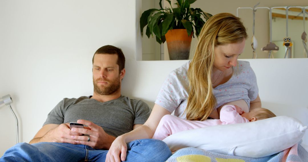 Mother Breastfeeding Baby While Father Uses Smartphone on Couch - Free Images, Stock Photos and Pictures on Pikwizard.com