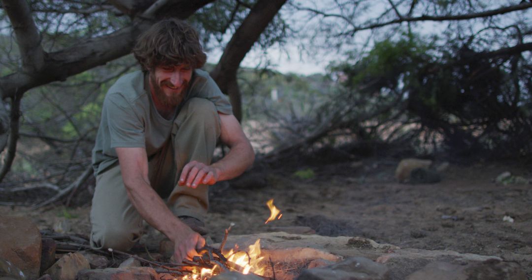 Happy bearded caucasian male survivalist adding kindling to fire at camp in wilderness - Free Images, Stock Photos and Pictures on Pikwizard.com