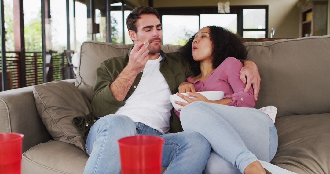 Smiling biracial couple eating popcorn sitting together on the couch at vacation home - Free Images, Stock Photos and Pictures on Pikwizard.com