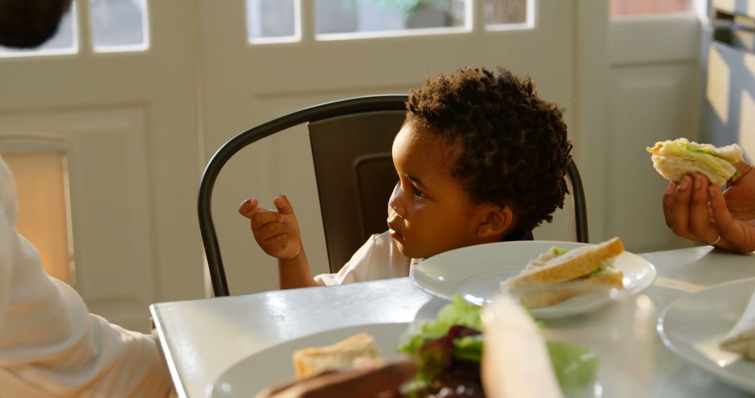 Child Eating Sandwich with Family in Sunlit Kitchen - Free Images, Stock Photos and Pictures on Pikwizard.com