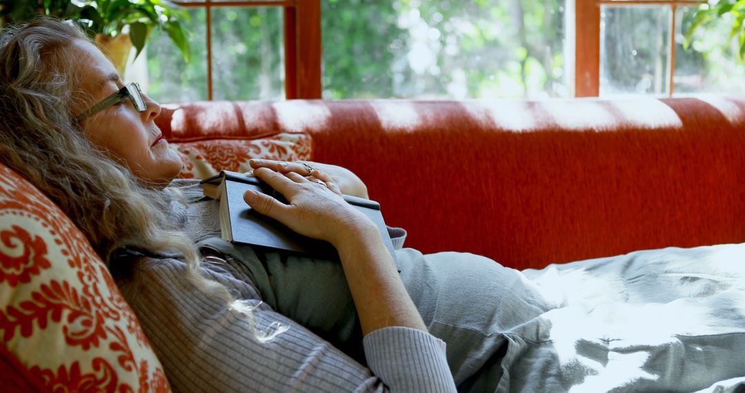 Senior Woman Resting on Sofa with Book in Sunlit Room - Free Images, Stock Photos and Pictures on Pikwizard.com