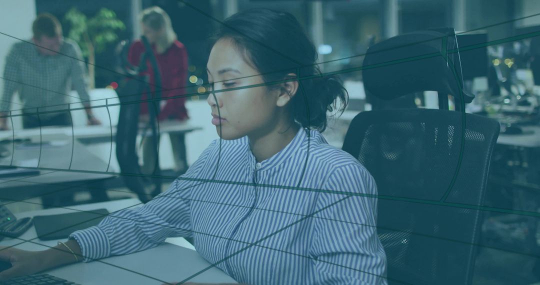 Focused woman typing in modern open-plan office with glass reflections and teamwork