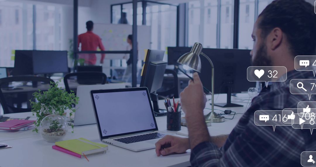 Bearded man typing on laptop and smartphone in open-plan office with social media overlays
