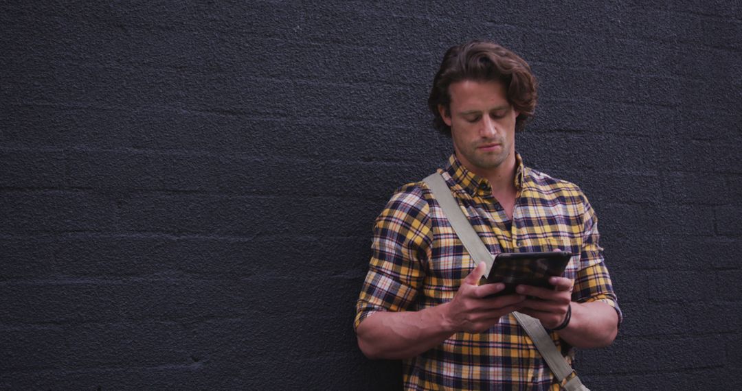 Young Man in Plaid Shirt Using Tablet Against Dark Brick Wall - Free Images, Stock Photos and Pictures on Pikwizard.com