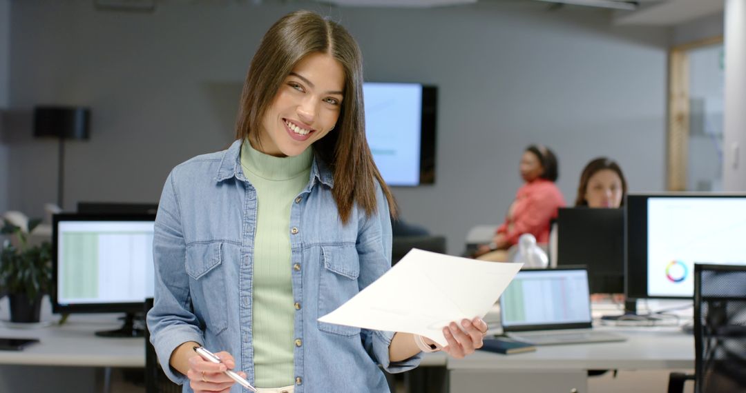 Smiling Indian woman holding documents and pen in bright modern open office environment