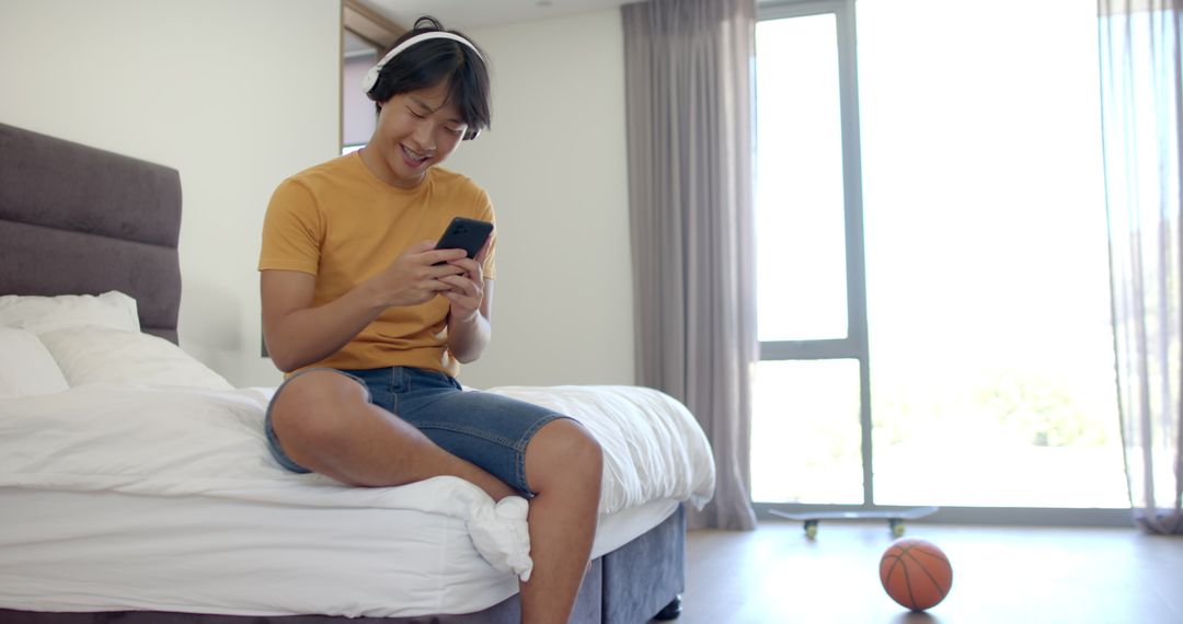 Young Asian Man Relaxing at Home Using Smartphone and Headphones