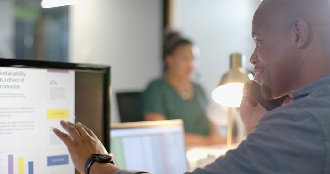 African American professional talking on phone pointing at monitor in open-plan office