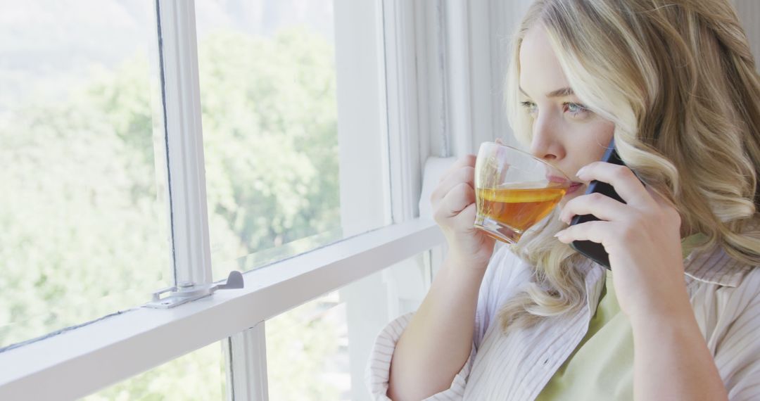 Young Woman Drinking Tea and Talking on Phone by Sunny Window - Free Images, Stock Photos and Pictures on Pikwizard.com