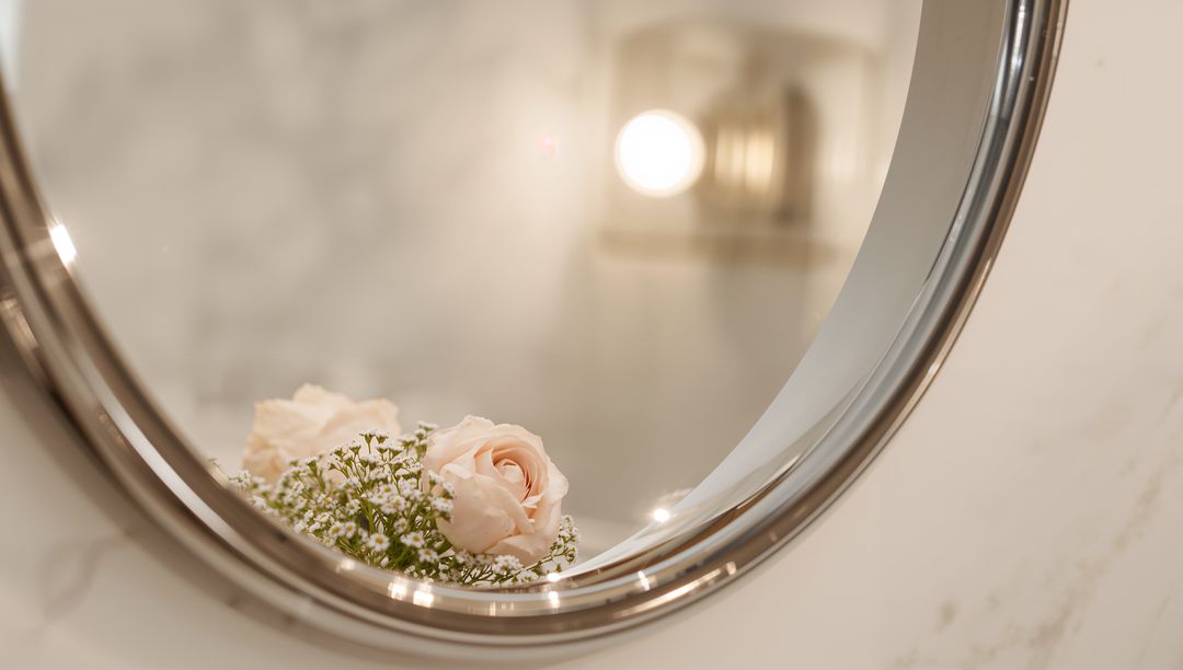Blush roses reflecting in round metal mirror over marble vanity with soft sconce glow