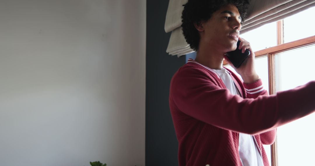 African American man talking on phone while adjusting window blind in minimalist interior