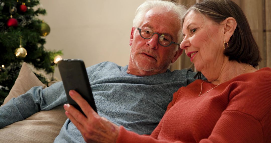 Senior Couple Sharing Tablet on Sofa with Christmas Tree in Background