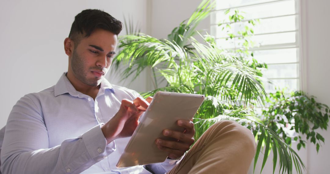 Young Man Working on Digital Tablet in Bright Modern Room with Green Plants - Free Images, Stock Photos and Pictures on Pikwizard.com