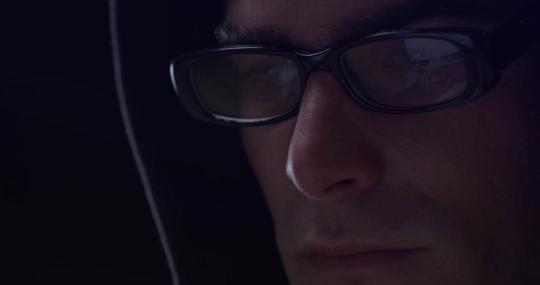 Hooded Man with Reflection on Glasses Focused on Screen in Dark Room