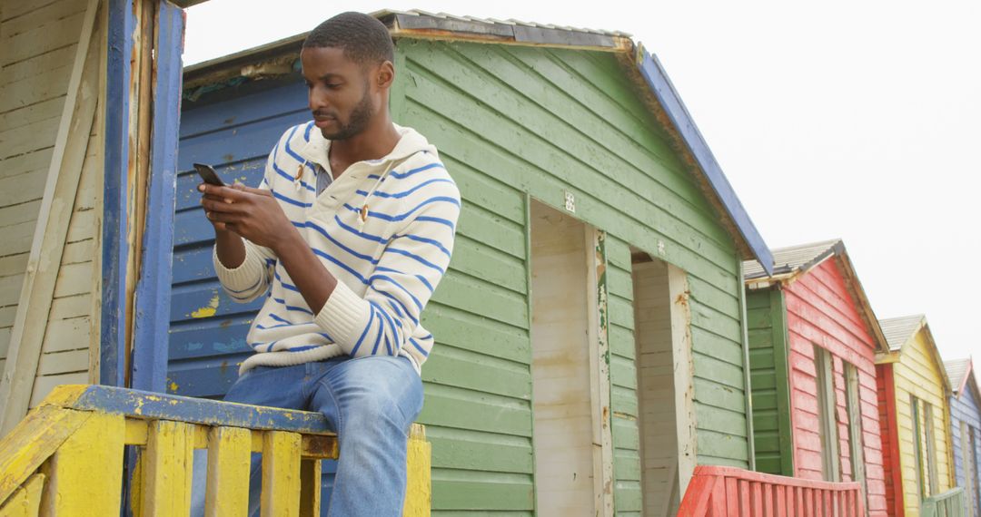 Casual Young Man Checking Smartphone by Colorful Beach Huts - Free Images, Stock Photos and Pictures on Pikwizard.com