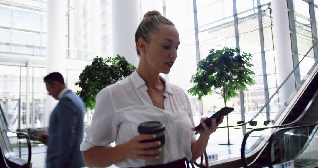 Businesswoman using smartphone while holding coffee cup in modern office lobby - Free Images, Stock Photos and Pictures on Pikwizard.com