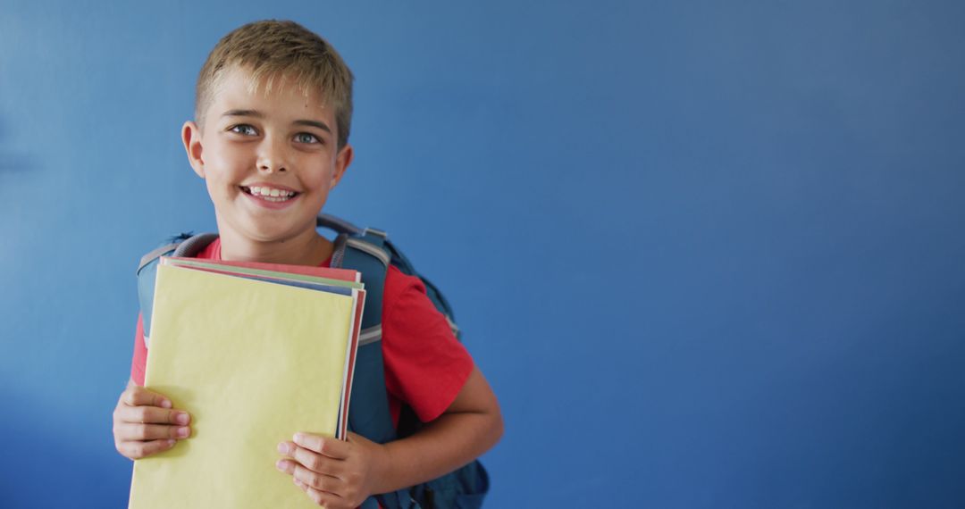 Happy Schoolboy with Backpack Holding Notebooks Against Blue Wall - Free Images, Stock Photos and Pictures on Pikwizard.com