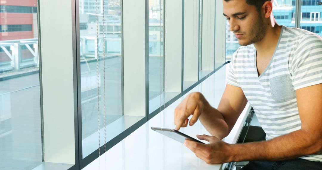 Young man using a tablet by modern office window - Free Images, Stock Photos and Pictures on Pikwizard.com