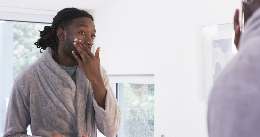African American man applying moisturizer at mirror in gray bathrobe morning routine