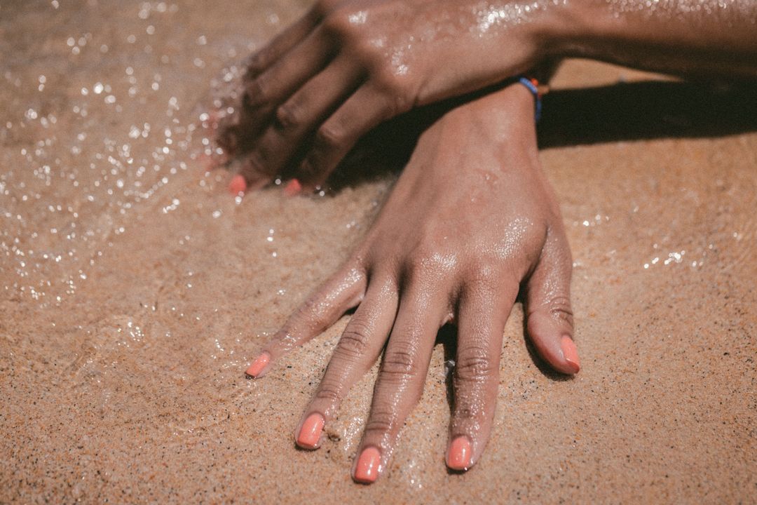 Sunlit wet hands resting on golden sand showing coral nail polish and beach bracelet