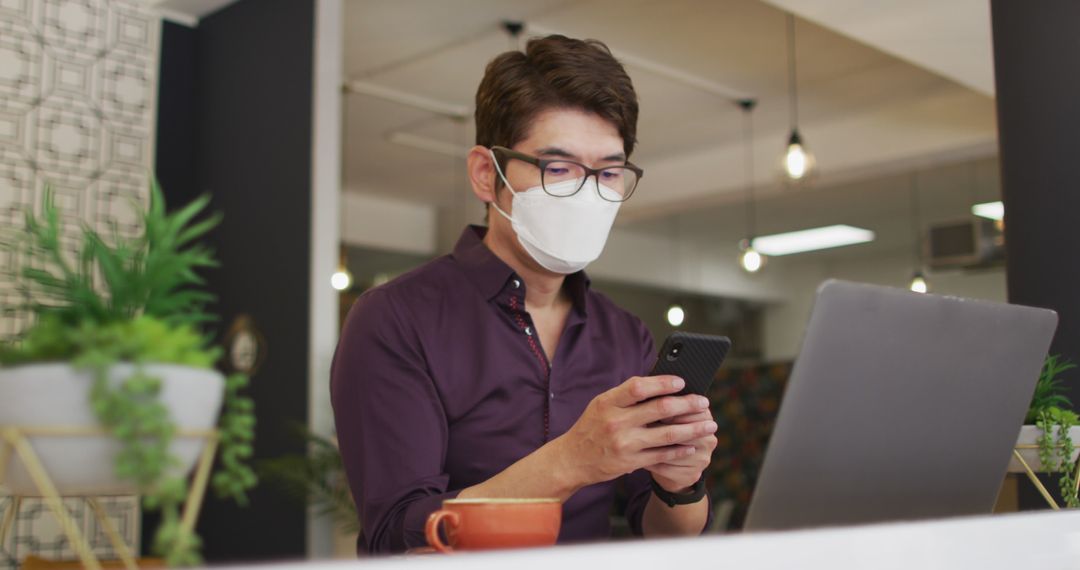 Young Asian professional wearing face mask using smartphone and laptop in modern office - Free Images, Stock Photos and Pictures on Pikwizard.com