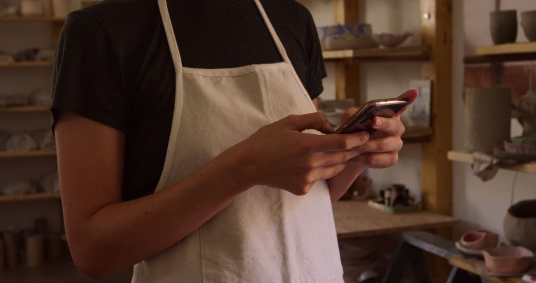 Person Wearing Apron Using Smartphone in Cozy Pottery Studio - Free Images, Stock Photos and Pictures on Pikwizard.com