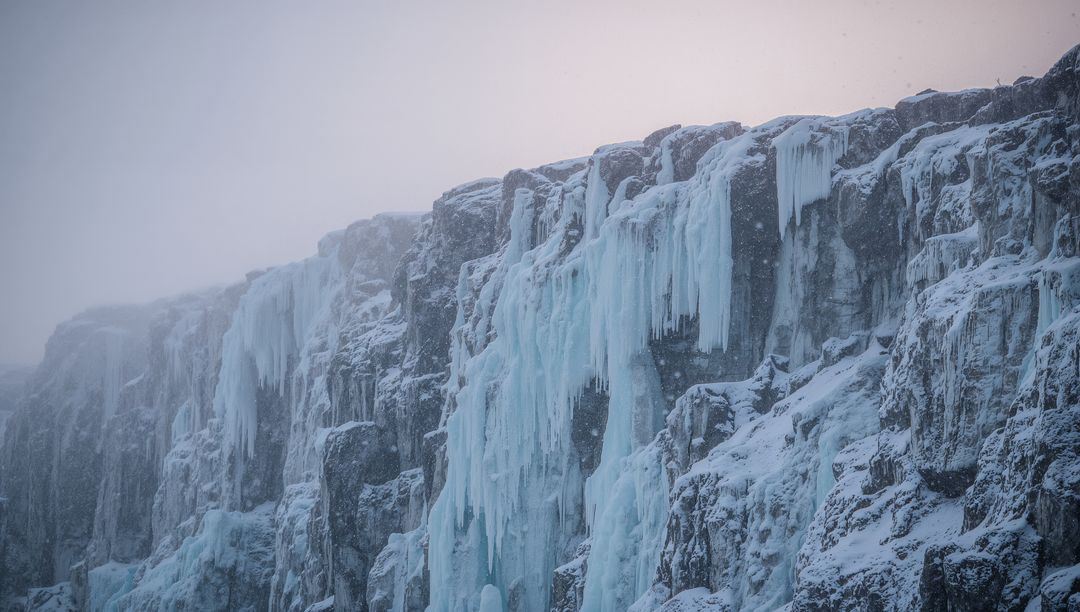 Frozen cliff draping cascading turquoise ice and icicles, snow falling through mist