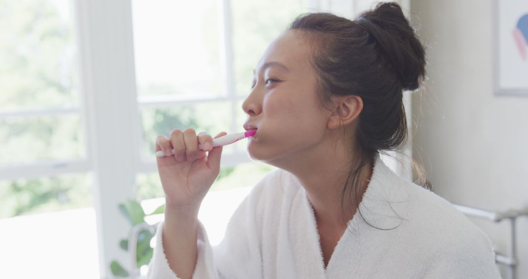 Young woman brushing teeth in bathroom wearing white robe - Free Images, Stock Photos and Pictures on Pikwizard.com