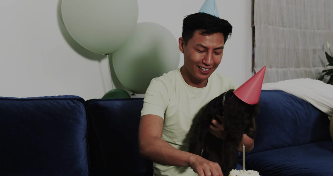 Smiling man celebrating birthday with dog on couch wearing party hats and cutting cake