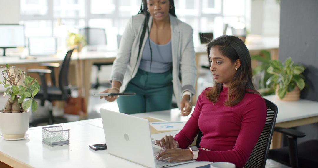 Multicultural female colleagues collaborating at open-plan office using laptop and tablet