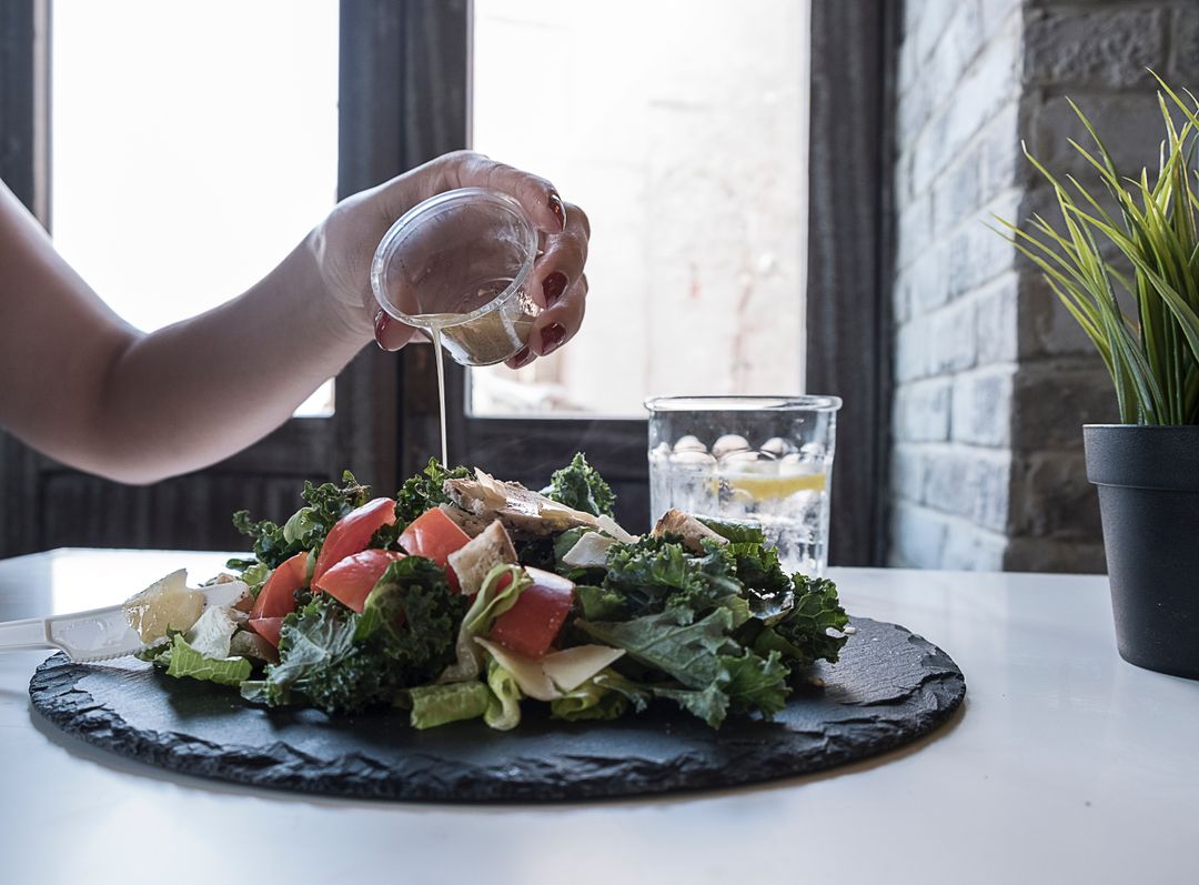 Pouring Lemon Vinaigrette Over Kale and Tomato Salad on Slate Plate Near Window