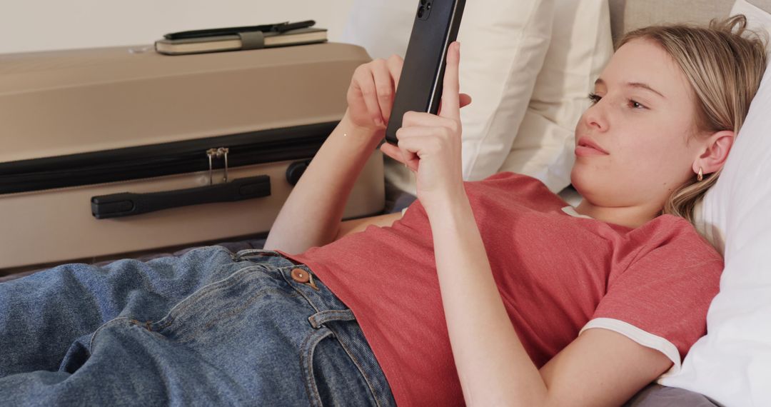 Young woman relaxing on bed scrolling smartphone beside packed suitcase and notebook