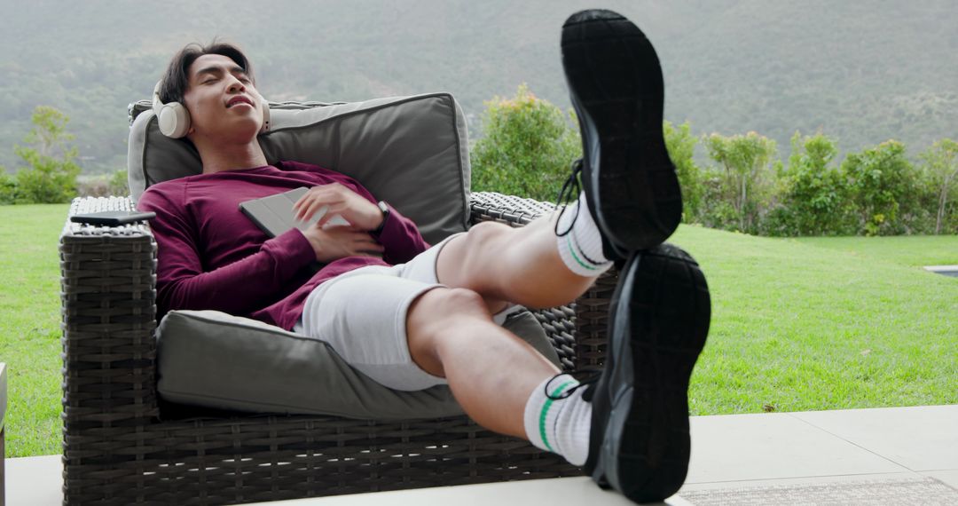 Asian Man Relaxing on Patio Lounger with Headphones and Tablet
