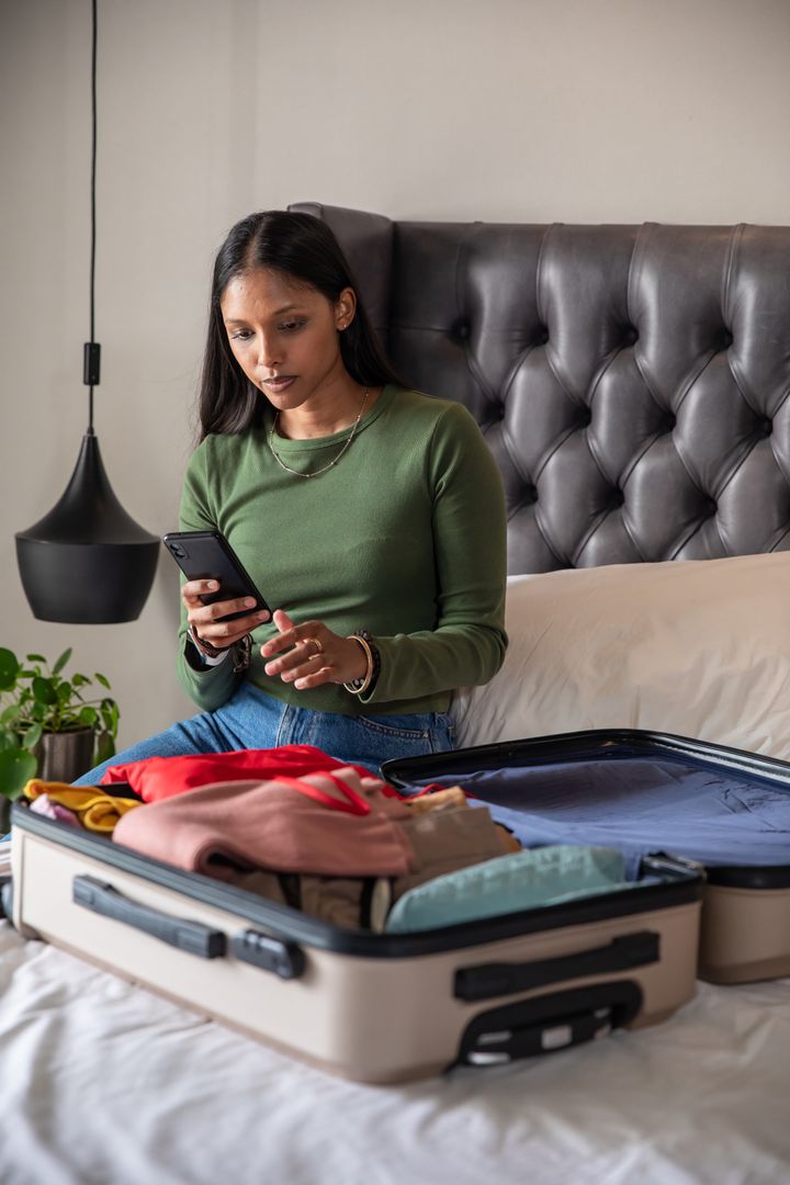 Woman Planning Travel and Packing Suitcase While Using Phone at Home