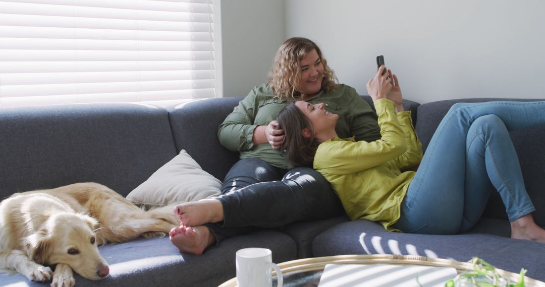 Caucasian lesbian couple smiling and sitting on couch with dog - Free Images, Stock Photos and Pictures on Pikwizard.com