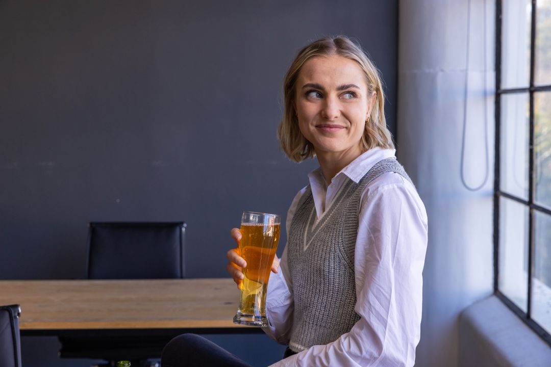 Smiling Woman Enjoying Beer by Window in Cozy Workplace