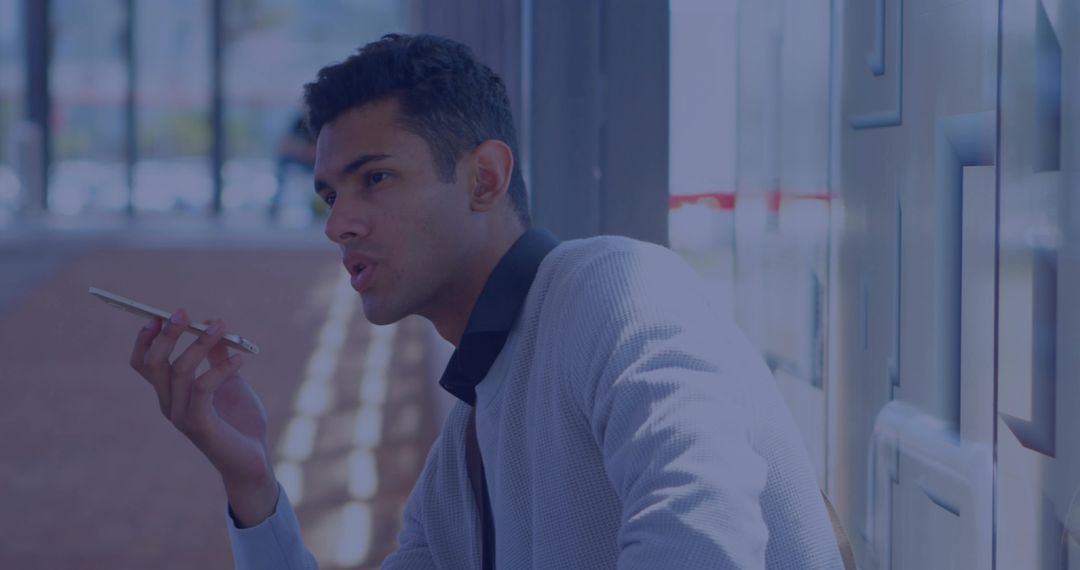 Indian professional speaking into smartphone by window in corridor wearing gray jacket