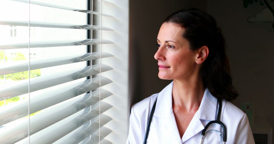 Female Doctor Looking Through Window with Contemplative Expression - Free Images, Stock Photos and Pictures on Pikwizard.com