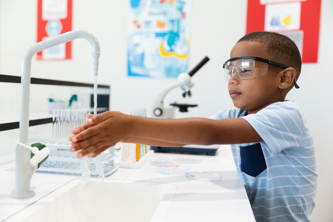 African american elementary schoolboy wearing protective eyewear washing hands in laboratory - Free Images, Stock Photos and Pictures on Pikwizard.com