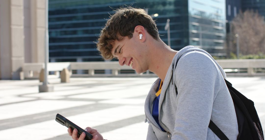Young man smiling while checking smartphone, wearing earbuds and backpack in urban plaza