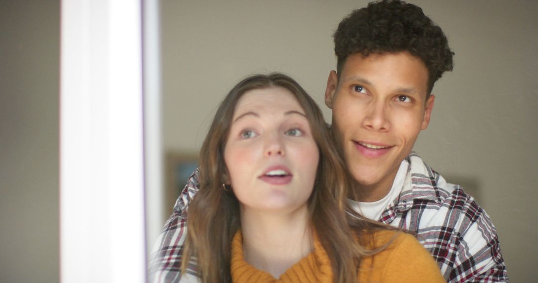 Happy Diverse Couple Embracing and Looking Through Window at Home