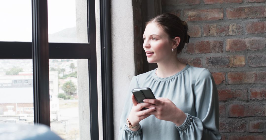 Young Professional Using Smartphone Near Window in Relaxed Office Setting