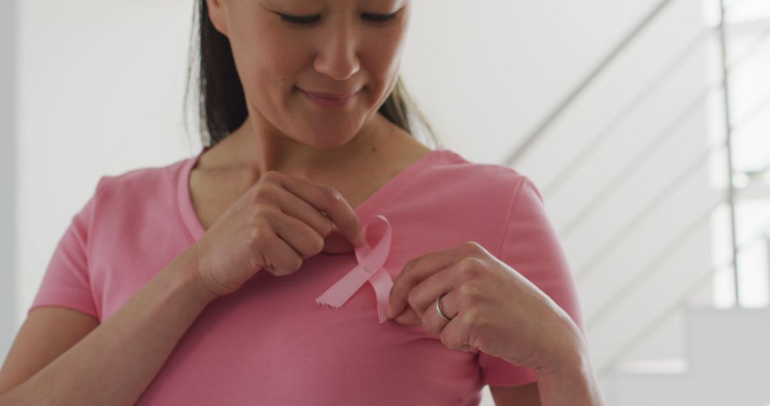 Happy asian woman in pink tshirt showing pink ribbon at home - Free Images, Stock Photos and Pictures on Pikwizard.com