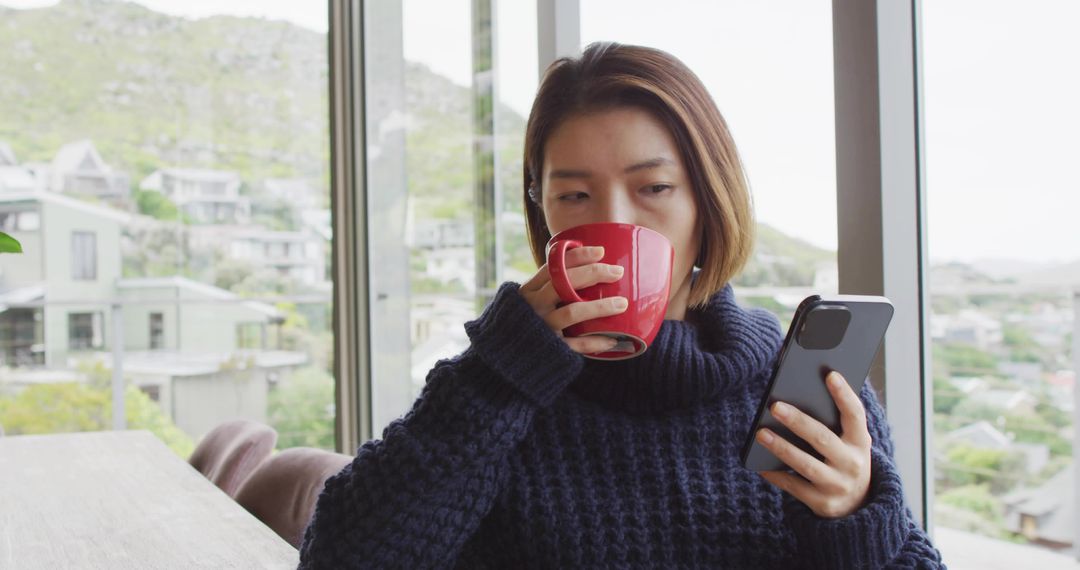 Sipping coffee while checking smartphone by window in cozy navy sweater, minimalist interior