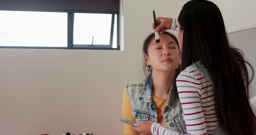 Friends applying makeup in bedroom using brushes and palettes, casual beauty routine