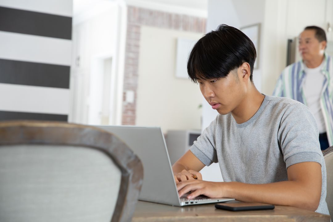 Focused Young Man Using Laptop at Home with Older Man Nearby