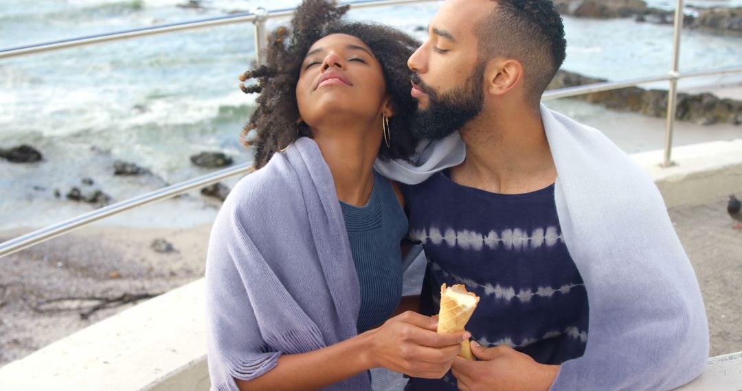 Romantic diverse couple with blankets on backs and ice creams embracing on sunny beach - Free Images, Stock Photos and Pictures on Pikwizard.com