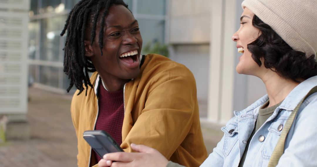 Laughing diverse friends sharing smartphone on urban bench near modern architecture