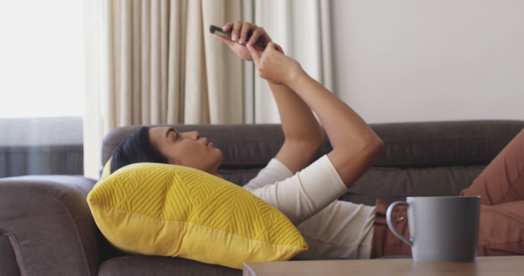 Young woman using smartphone while relaxing on couch with cup of coffee - Free Images, Stock Photos and Pictures on Pikwizard.com